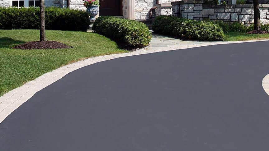 A freshly paved asphalt driveway curves toward a house, bordered by green grass and trimmed bushes, with touches of concrete along the edges.