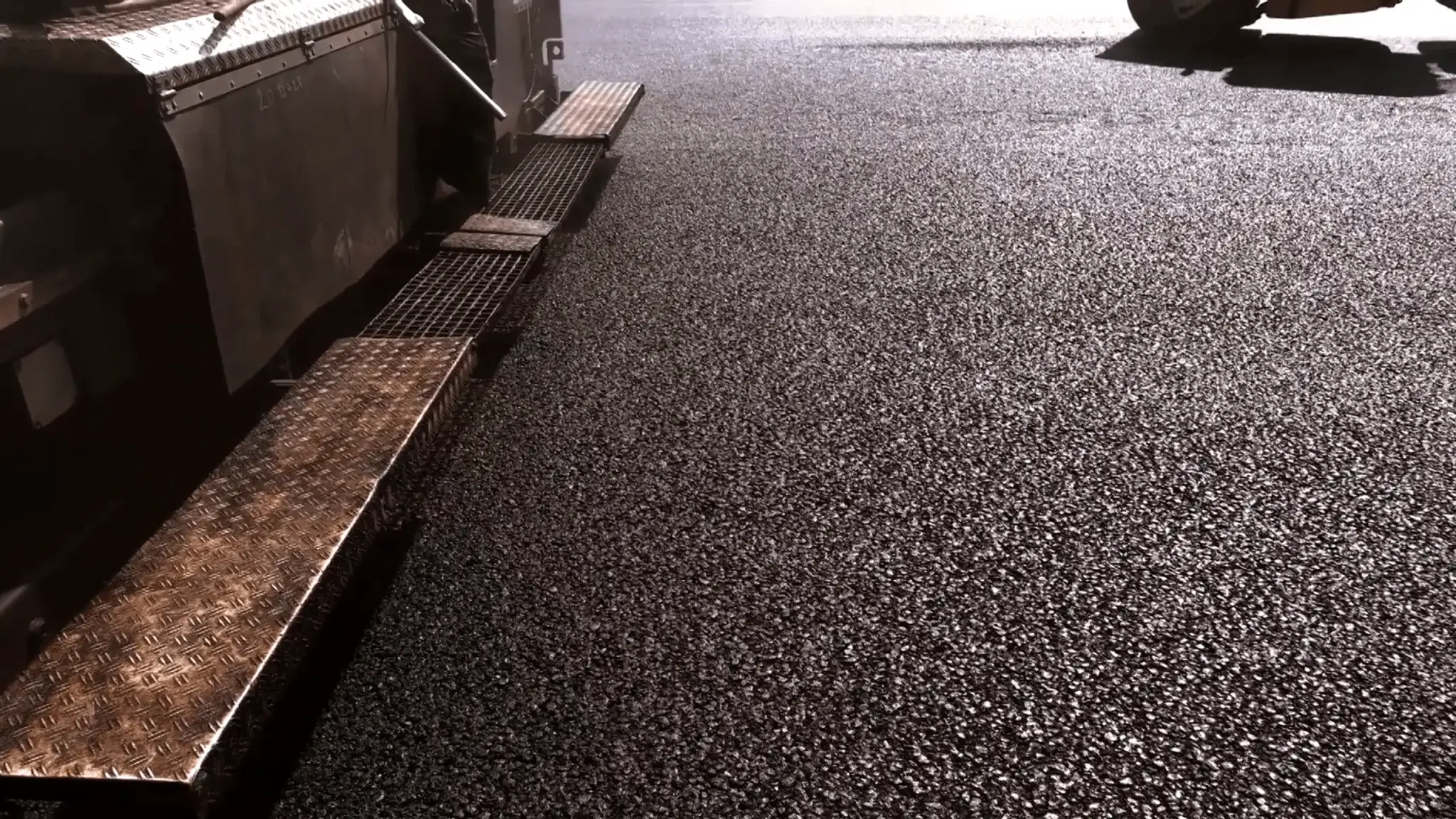 Close-up of fresh asphalt being laid by a paving machine on a road, with part of the machinery visible—a reminder of the importance to maintain your asphalt for long-lasting, smooth surfaces.