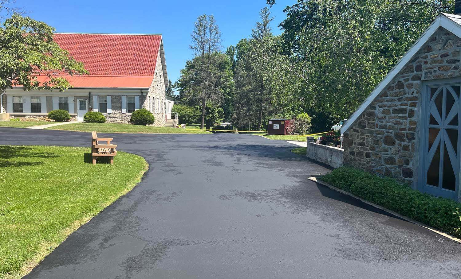 Paved driveway with expert driveway paving, stone buildings, green grass, and a wooden bench under a clear blue sky.