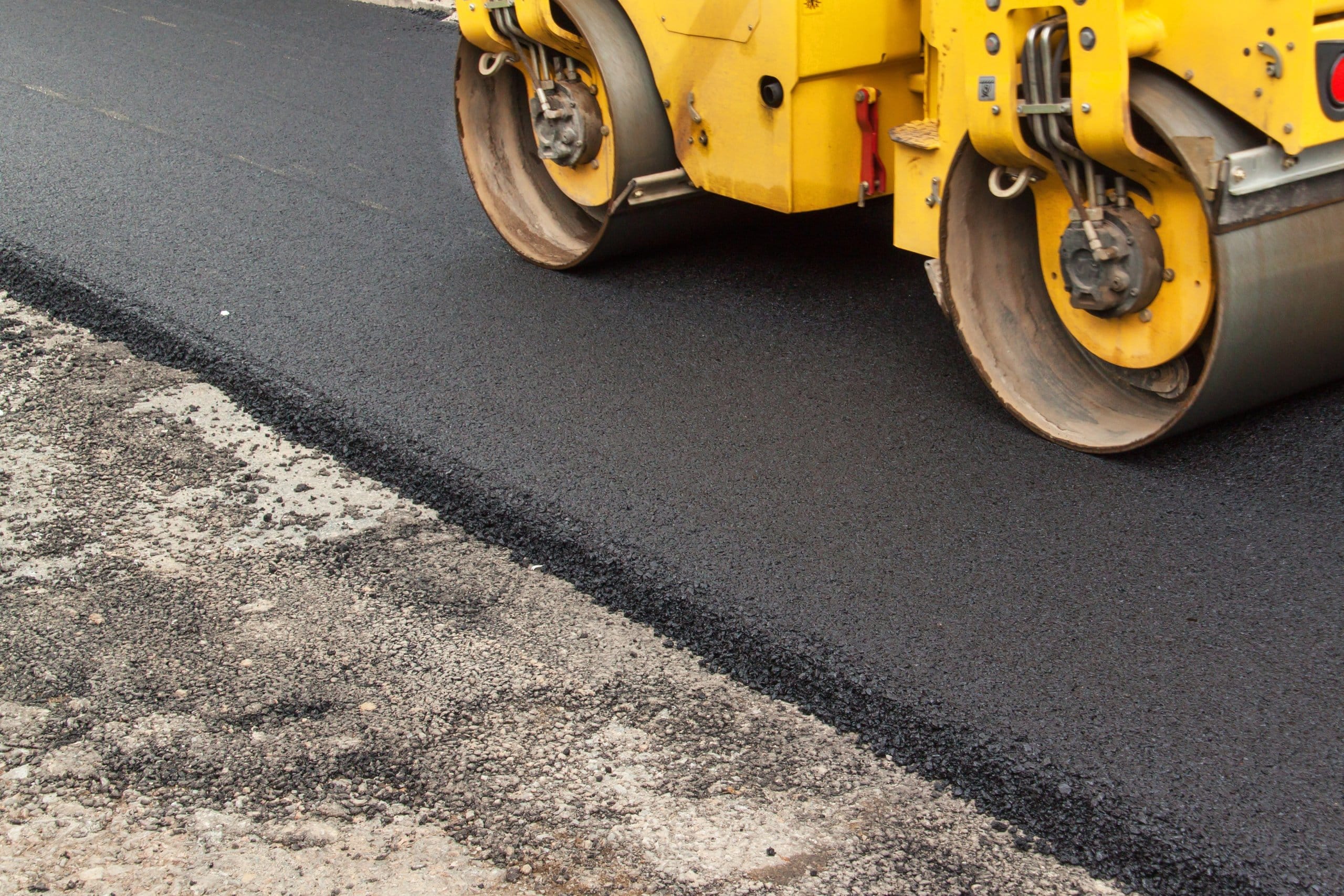 A yellow steamroller compacts a newly laid asphalt road surface. Concrete overlay.