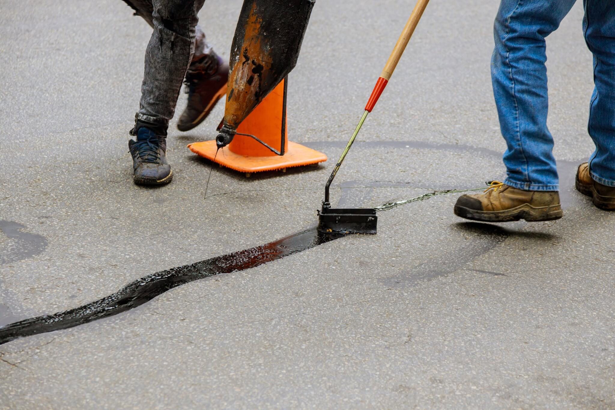 Road surface restoration work in the worker performs on road patcher work on the repair of cracks by filling and sealing with coated by bitumen emulsion Is Your Asphalt Cracking? Here’s Why Regular Sealcoating Is a Game-Changer Workers sealing a road crack with hot tar using a tool next to an orange cone.