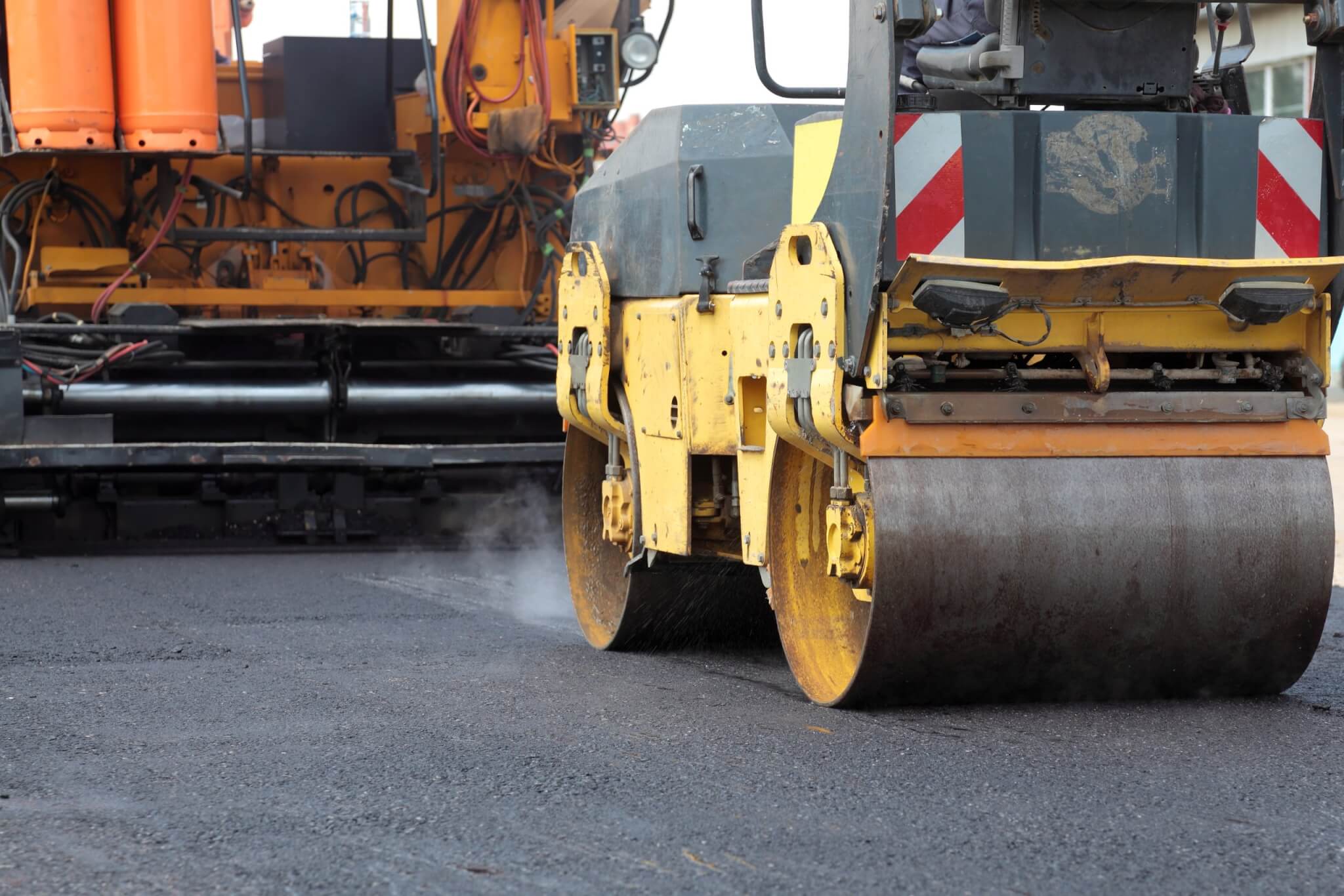 Roadwork road roller and paving machine at asphalt Yellow steamroller and a paver machine working on a fresh asphalt road, emitting steam.