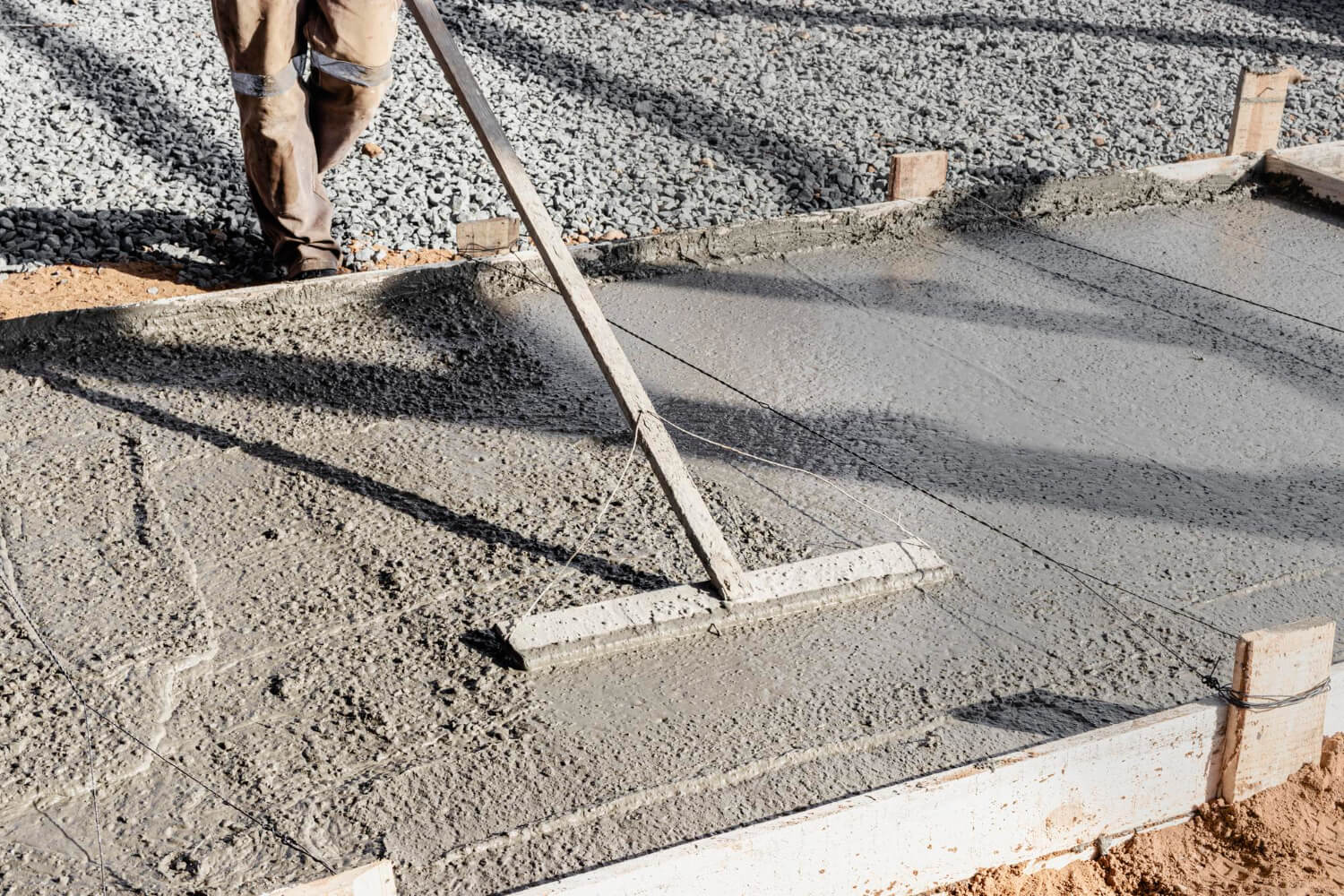 A worker smooths freshly poured concrete with a long-handled tool at a construction site.