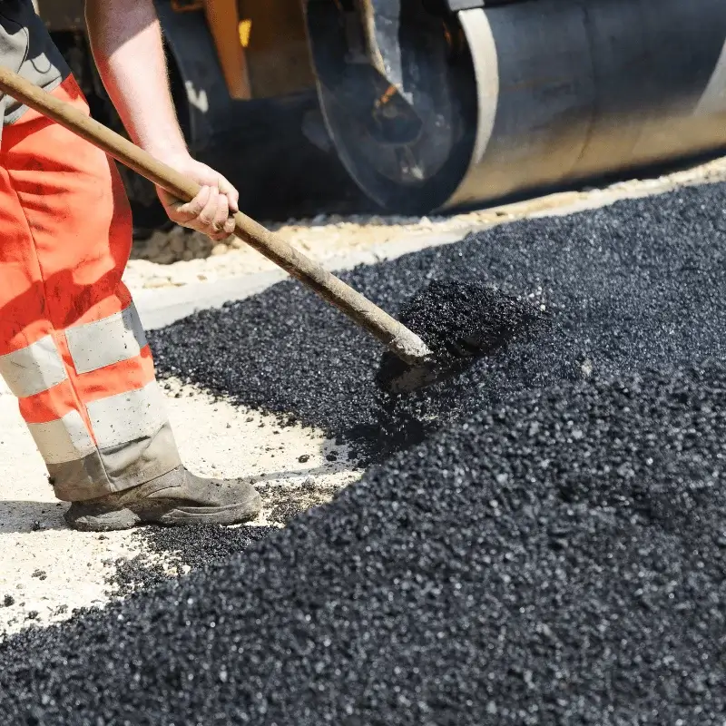 Worker in orange pants spreads asphalt with a shovel near a steamroller at a road construction site.