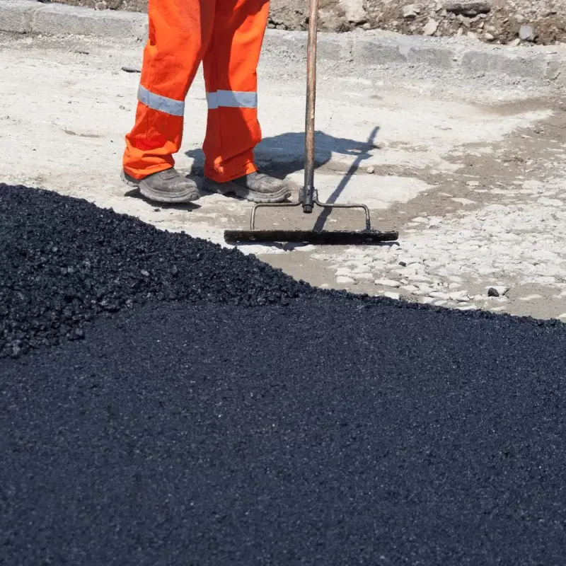 Worker in orange pants spreads fresh asphalt on a road with a rake.