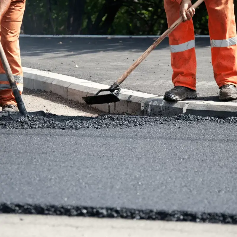 Two workers in orange pants level fresh asphalt on a road with shovels.
