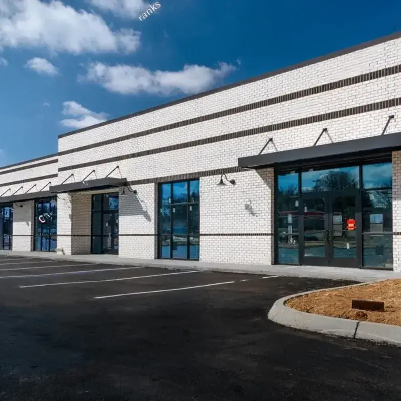 A modern, empty strip mall with white brick walls and large glass storefronts on a sunny day.