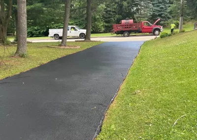 Freshly paved asphalt driveway with two work trucks and workers on a residential street surrounded by grass and trees.