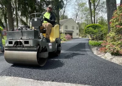 Worker operates steamroller smoothing fresh asphalt on a residential driveway, with another worker nearby.
