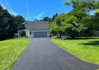 Paved driveway leading to a white garage, surrounded by green grass and trees on a sunny day.