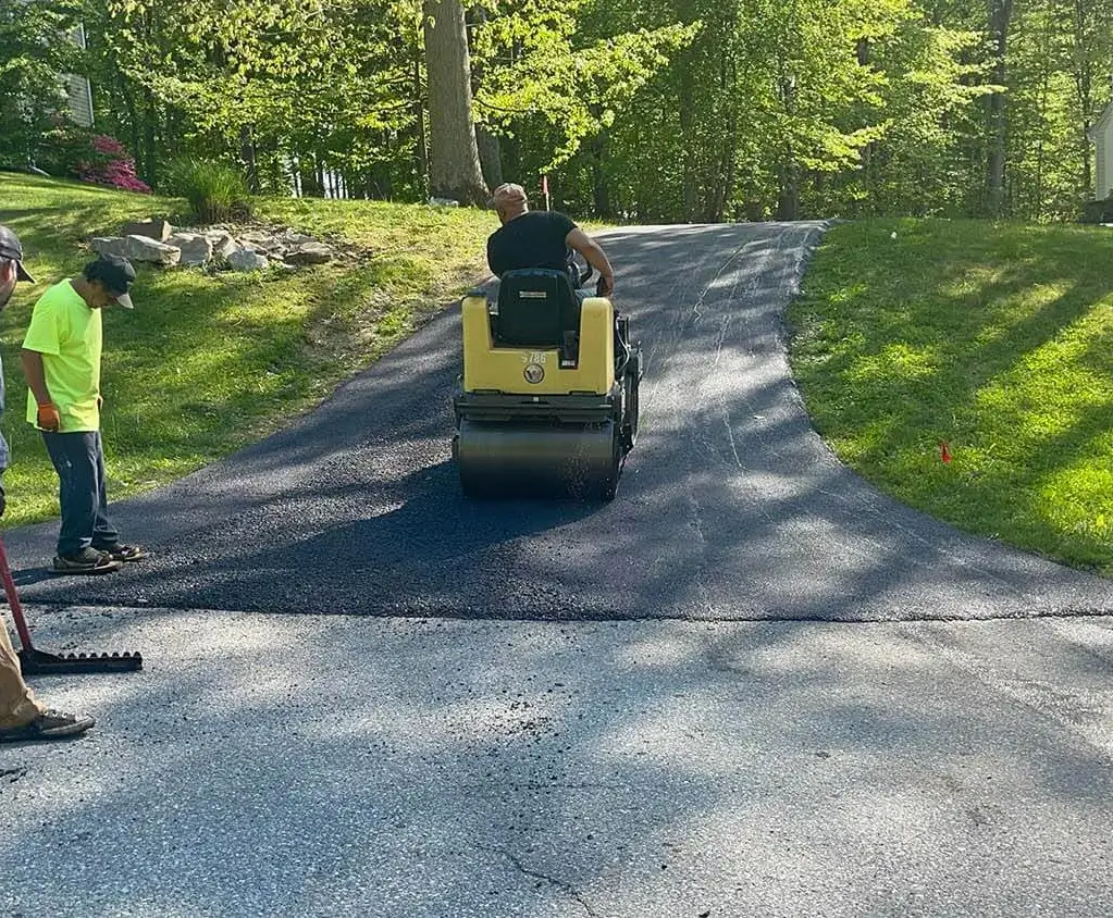 Workers paving a driveway with fresh asphalt; one operates a steamroller, others stand nearby with tools.