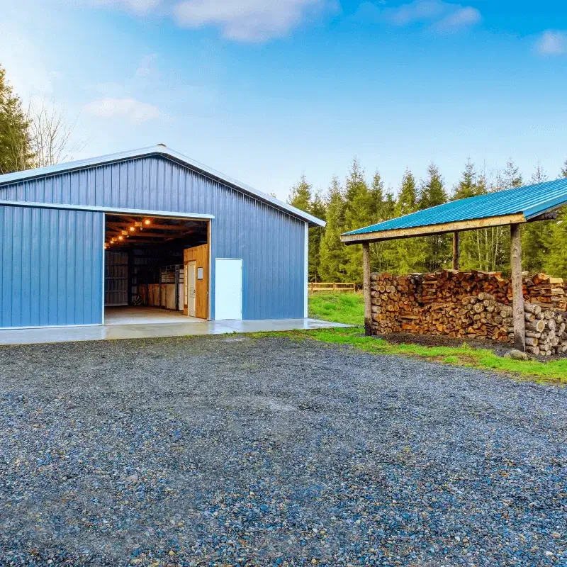 stone-gravel-2 A blue metal barn with its door open, next to a wood storage shed and gravel driveway.